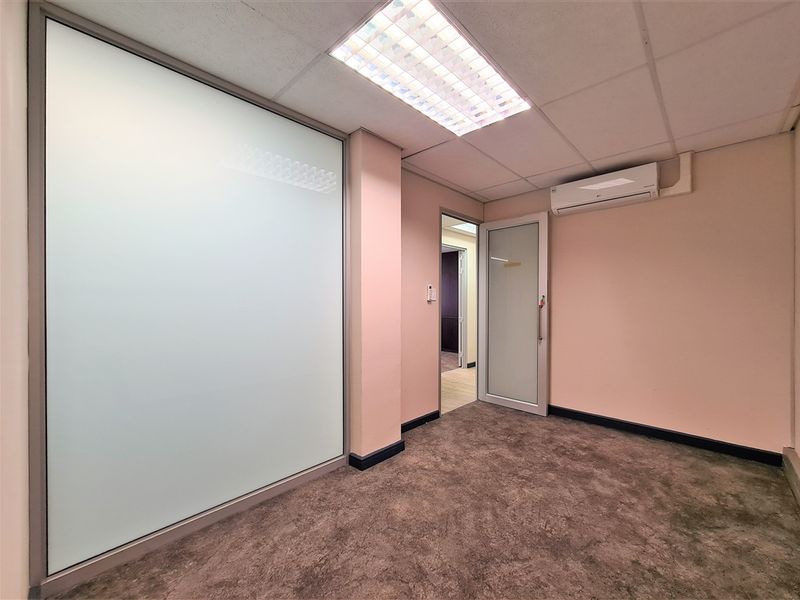 Empty office space featuring a frosted glass partition, wall-mounted air conditioning unit, suspended ceiling with recessed lighting, and carpeted flooring.