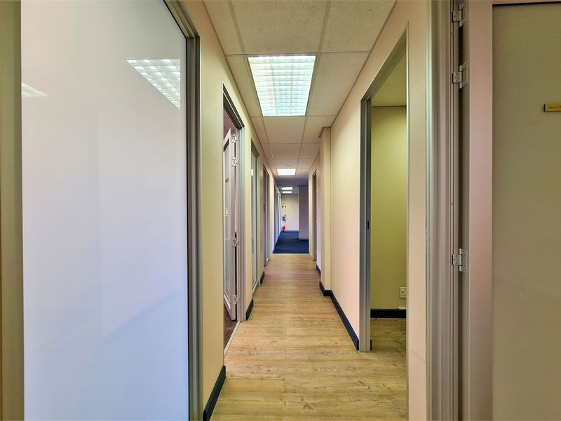 A clean, well-lit office hallway featuring wood-look flooring, suspended ceilings with recessed lighting, and multiple access points to individual office spaces.
