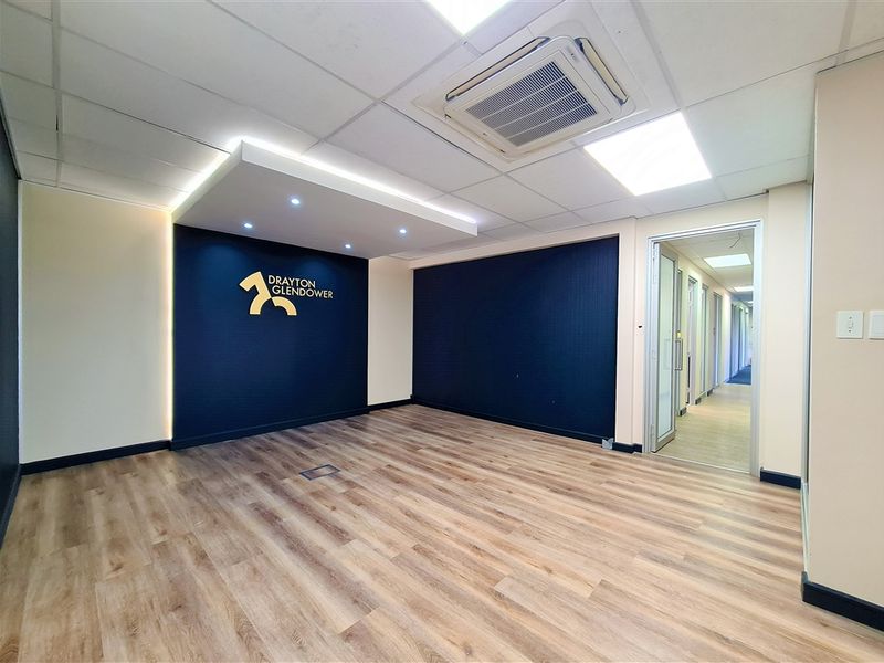 A modern, vacant office reception area featuring wood-look laminate flooring, a branded feature wall with accent lighting, and a suspended ceiling with integrated air conditioning.