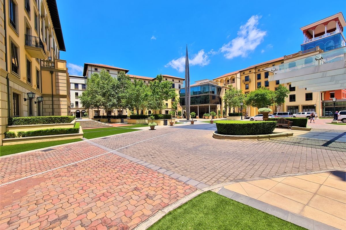 A well-maintained paved office courtyard featuring landscaped garden beds, artificial turf, and a central sculpture within a modern commercial complex.