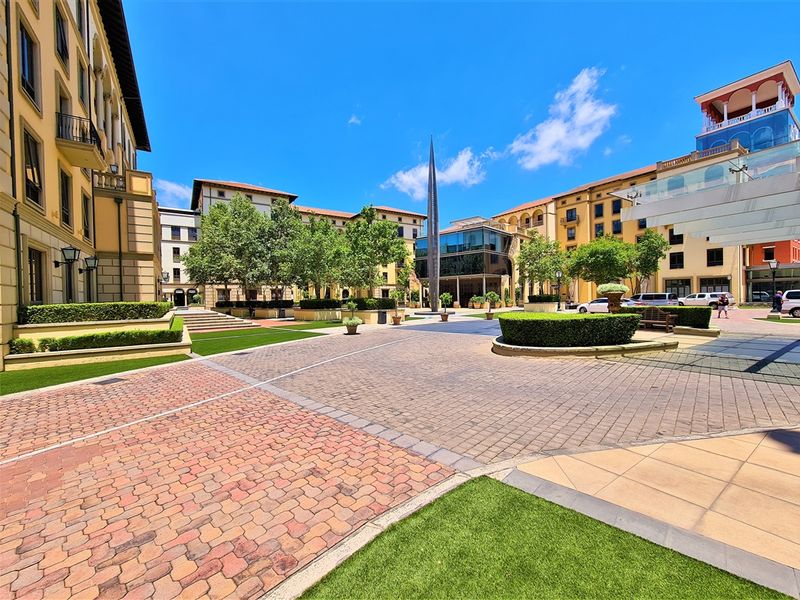 A well-maintained paved office courtyard featuring landscaped garden beds, artificial turf, and a central sculpture within a modern commercial complex.
