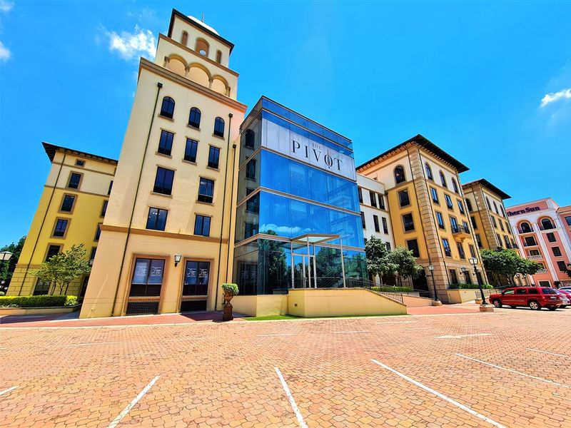 A modern office building facade featuring a prominent glass entrance tower and classic architectural elements, set against a paved parking area.
