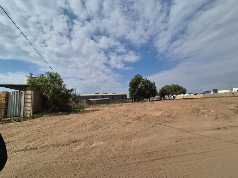 A wide-angle view of a vacant commercial land plot featuring a perimeter wall, metal gate, and existing silo structure.