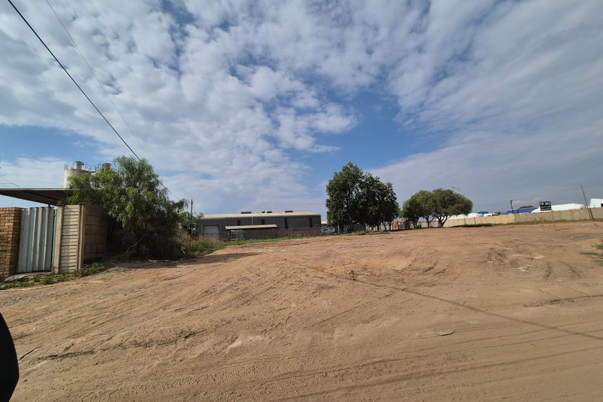 A wide-angle view of a vacant commercial land plot featuring a perimeter wall, metal gate, and existing silo structure.