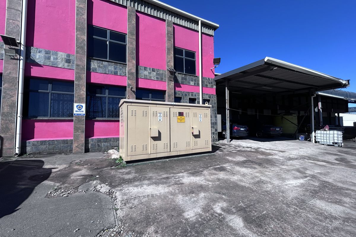 Industrial exterior view featuring a pink facade, large windows, a dedicated electrical substation unit, and a covered loading or parking area.