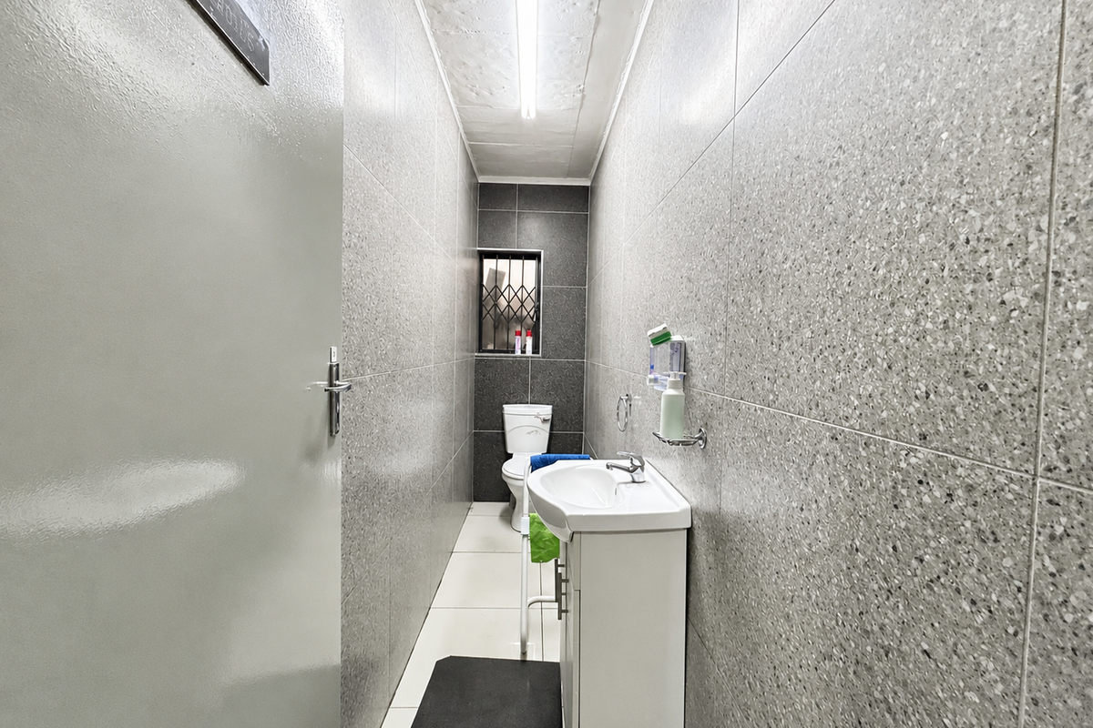 A compact, modern bathroom featuring full-height grey speckled wall tiling, a white vanity basin, and a toilet with a security-grilled window.