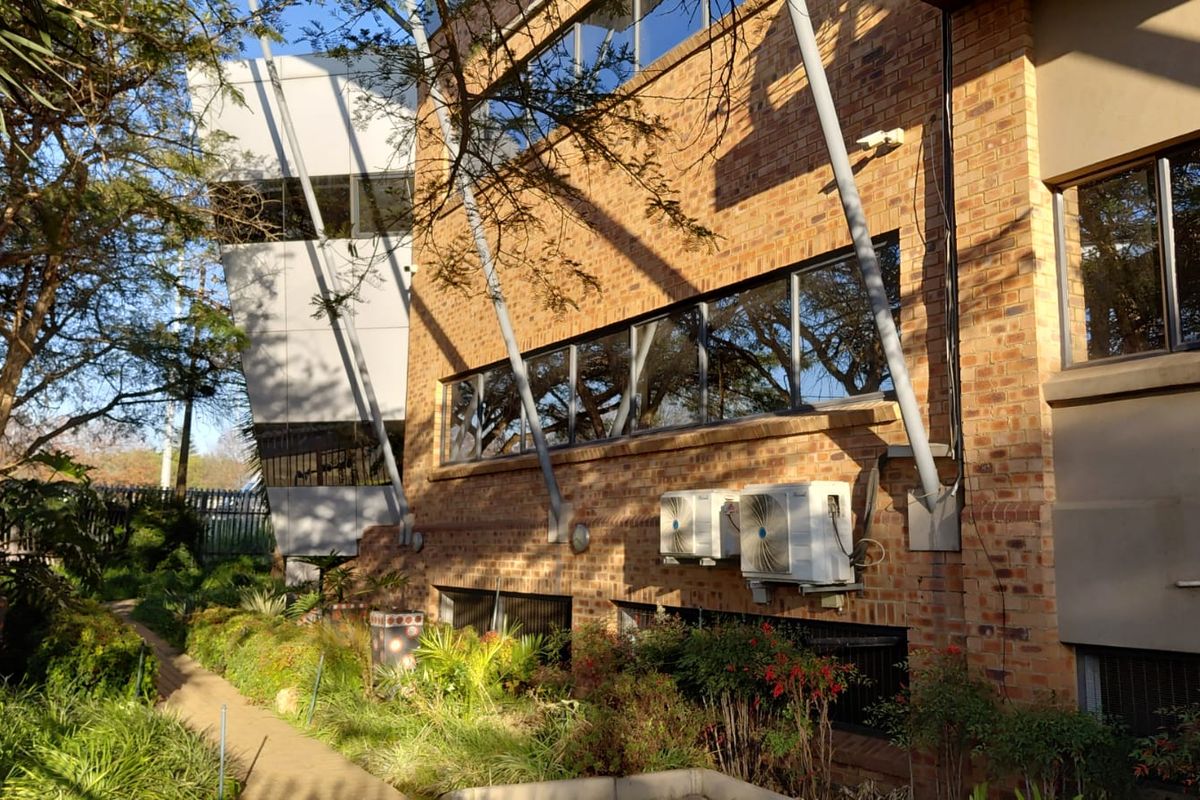 Exterior view of a brick commercial building featuring large windows, structural support beams, and wall-mounted air conditioning units set within a landscaped garden area.
