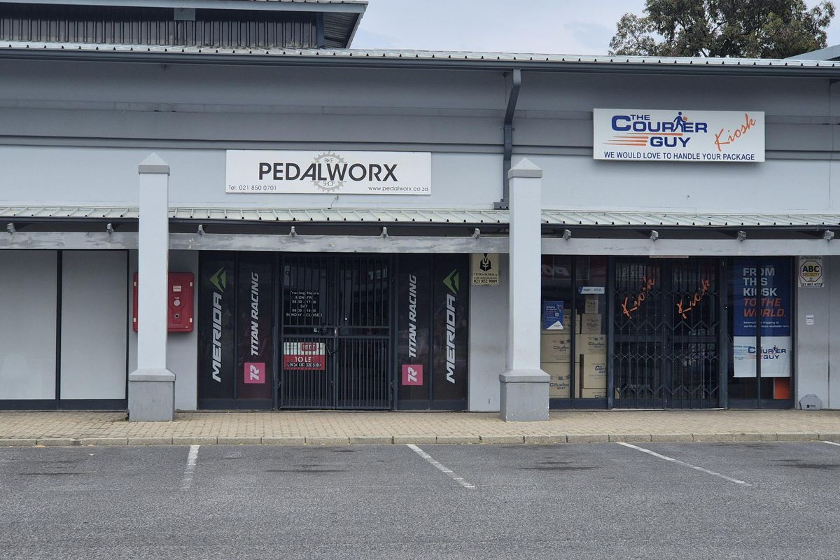 Front facade of a retail commercial property featuring dual storefronts with metal security gates, signage, and a paved parking area.