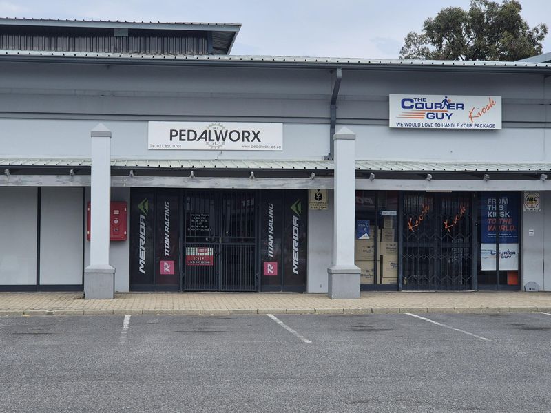 Front facade of a retail commercial property featuring dual storefronts with metal security gates, signage, and a paved parking area.