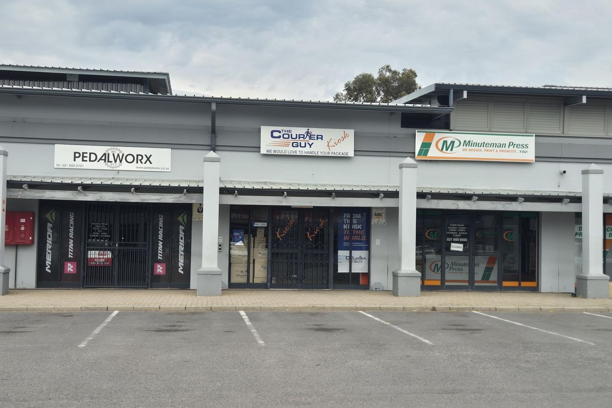 Front elevation of a retail strip center featuring multiple storefronts with signage, metal security gates, and a paved parking area.