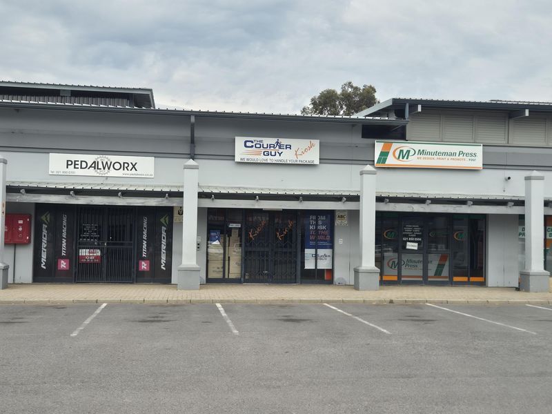 Front elevation of a retail strip center featuring multiple storefronts with signage, metal security gates, and a paved parking area.
