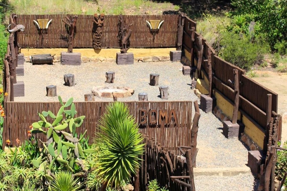 An outdoor boma area featuring a central fire pit, gravel flooring, and rustic wooden fencing, set within a landscaped garden environment.