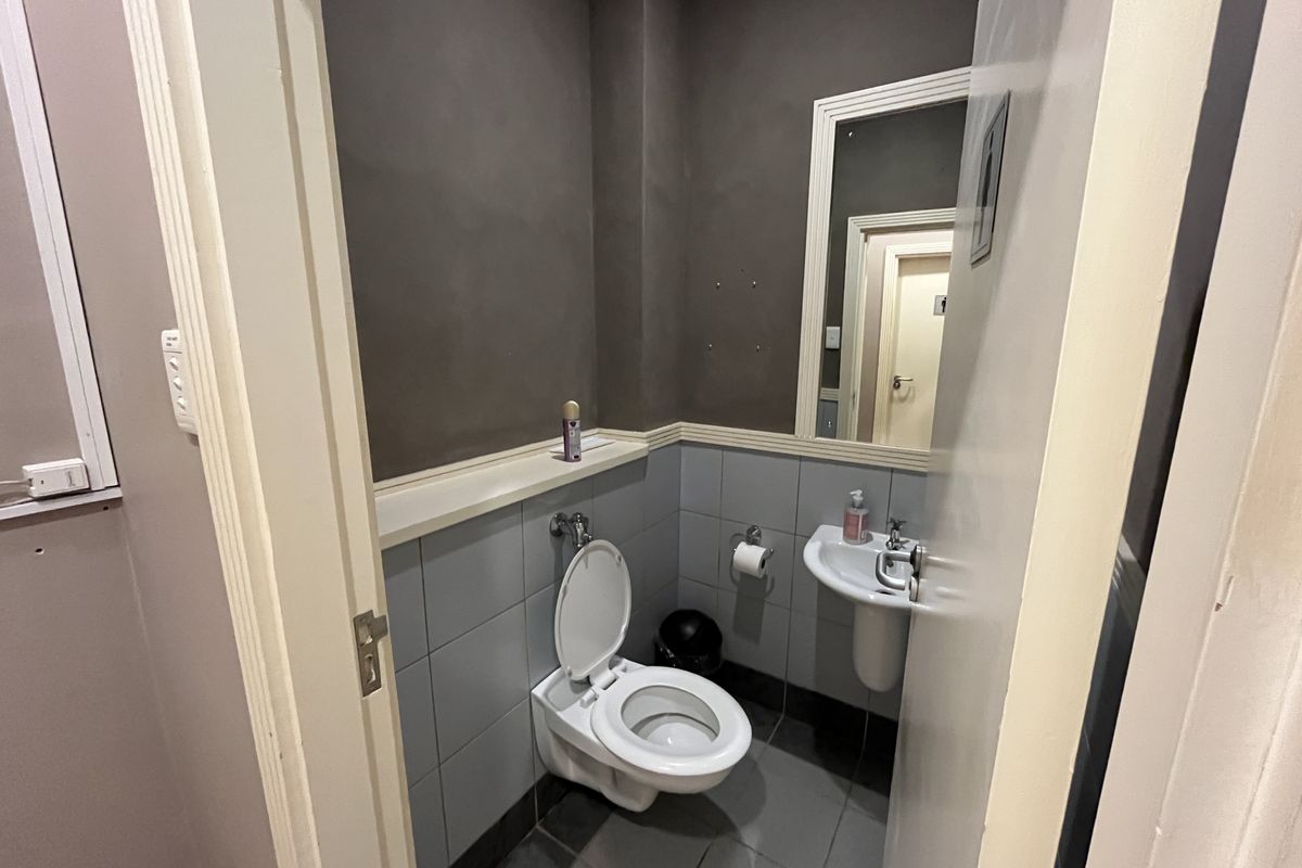A compact office restroom featuring grey wall tiling, a wall-mounted toilet, a small pedestal basin, and a framed mirror.