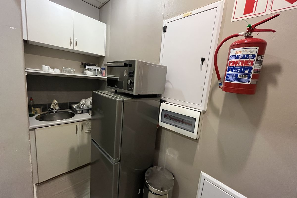 A compact office kitchenette featuring built-in cabinetry, a stainless steel sink, and essential safety equipment including a fire extinguisher and electrical distribution board.