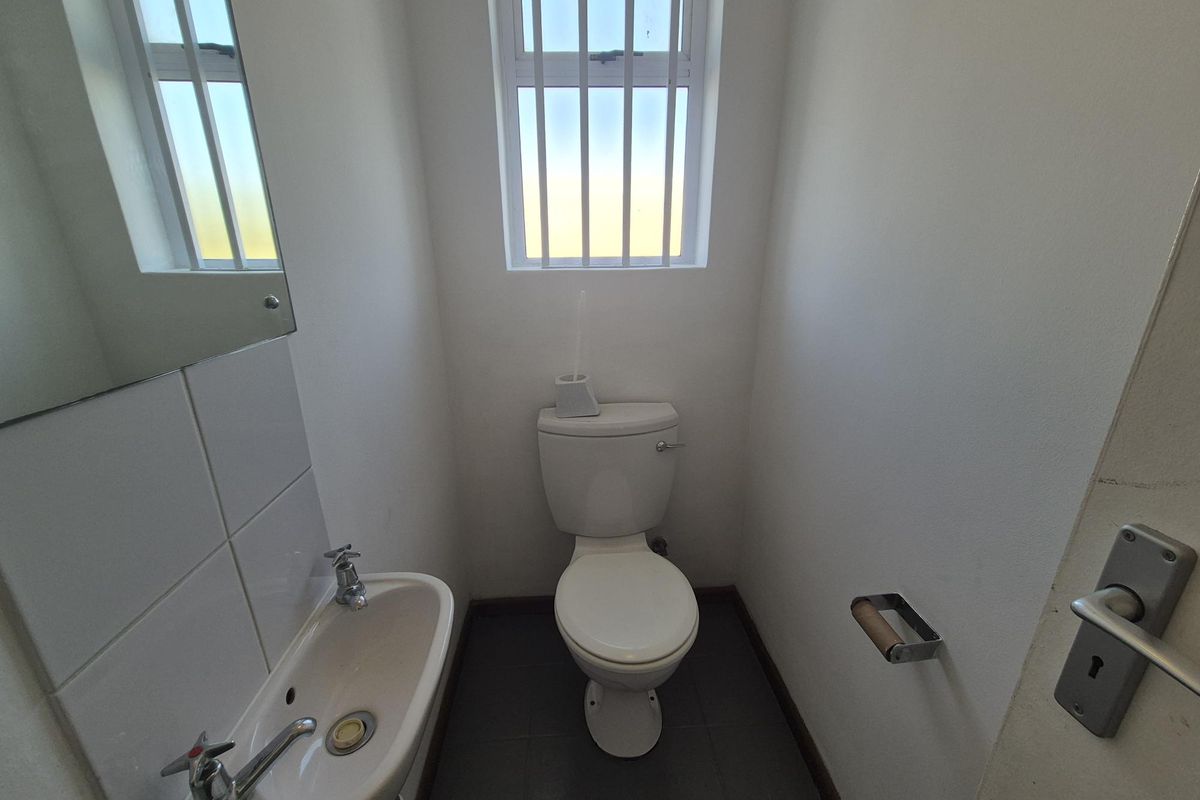 A compact industrial-style guest bathroom featuring a white toilet, wall-mounted basin with chrome taps, and a window with security bars.