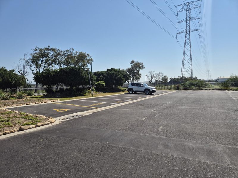 A paved industrial parking area featuring marked bays, including a designated disabled parking space, perimeter fencing, and established garden borders.