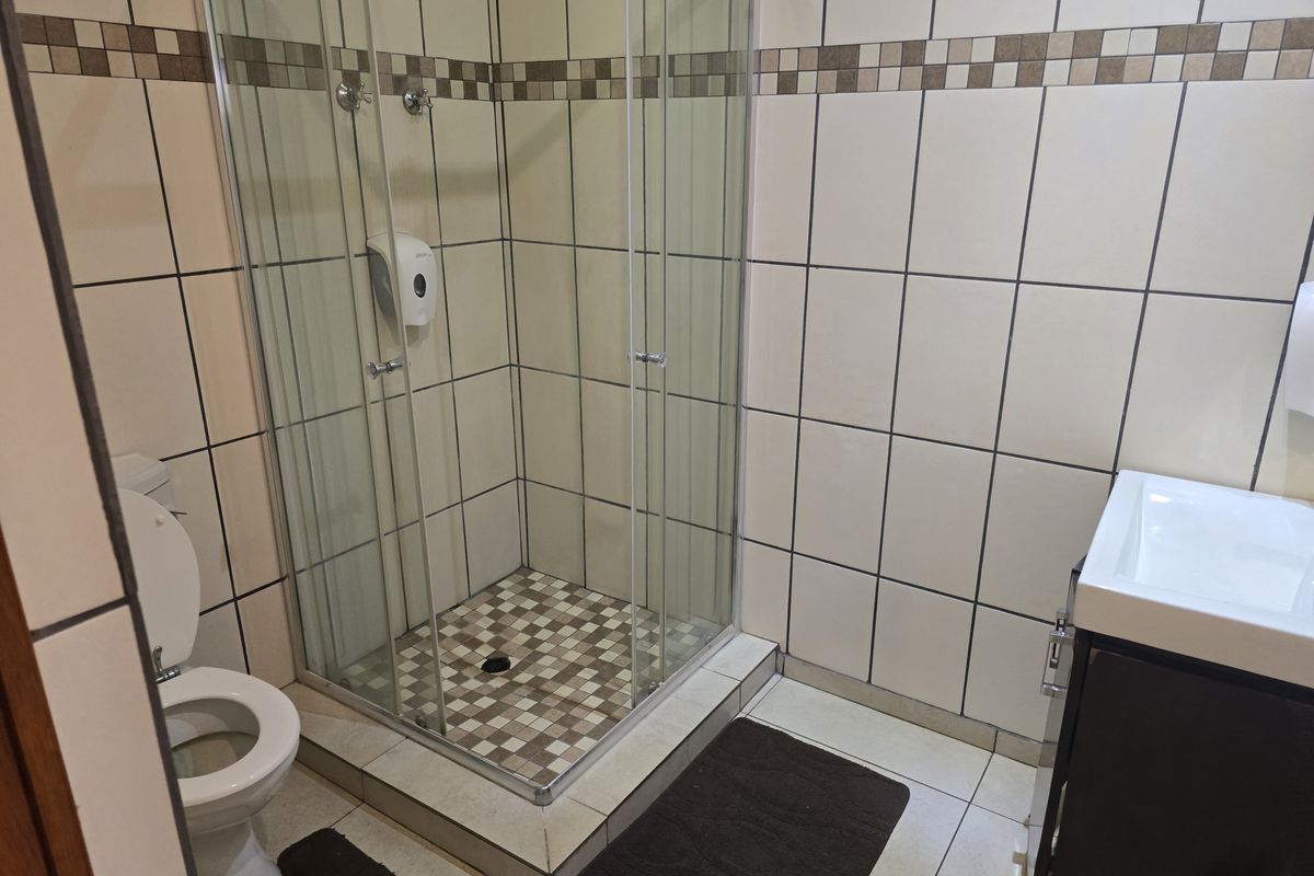 A functional hotel bathroom featuring a glass-enclosed shower, a wall-mounted toilet, and a vanity basin with tiled walls and flooring.