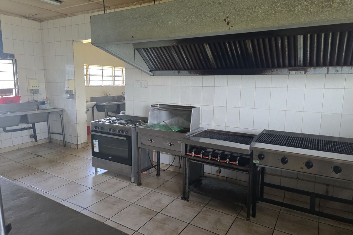 Commercial kitchen featuring a large stainless steel extractor hood, industrial gas stove, grill, and prep area with tiled walls and flooring.