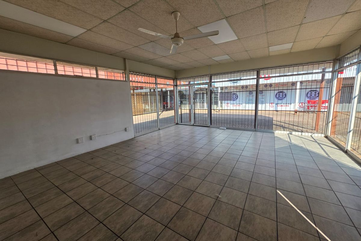 Vacant commercial office space featuring large glass storefront windows, security gates, ceramic tiled flooring, and a suspended ceiling with an integrated fan.