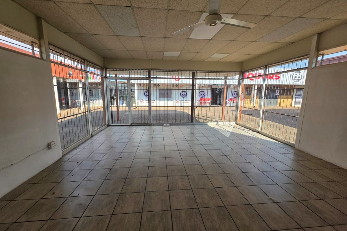 Empty commercial retail space featuring tiled flooring, a suspended ceiling with a fan, and large glass storefront windows with security bars.