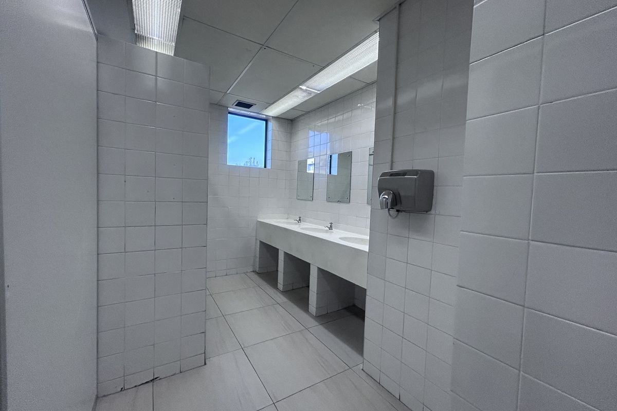 A functional commercial office restroom featuring a multi-basin vanity, tiled walls and flooring, and integrated fluorescent lighting.
