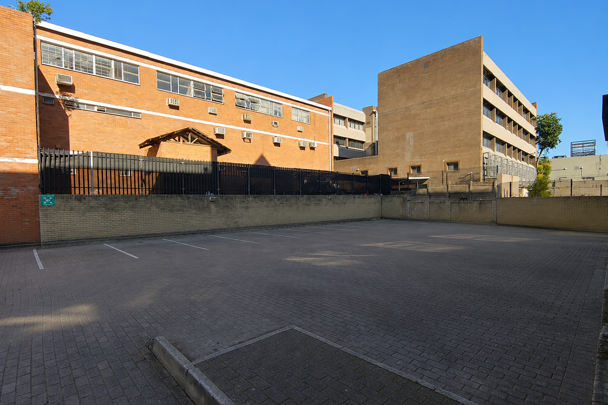 A paved outdoor parking area situated between office buildings, featuring brick boundary walls and perimeter security fencing.