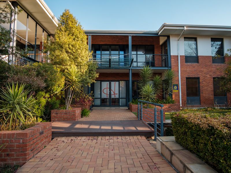Front elevation of a professional office building featuring a brick facade, glass entrance, balcony, and landscaped paved walkway with a timber footbridge.