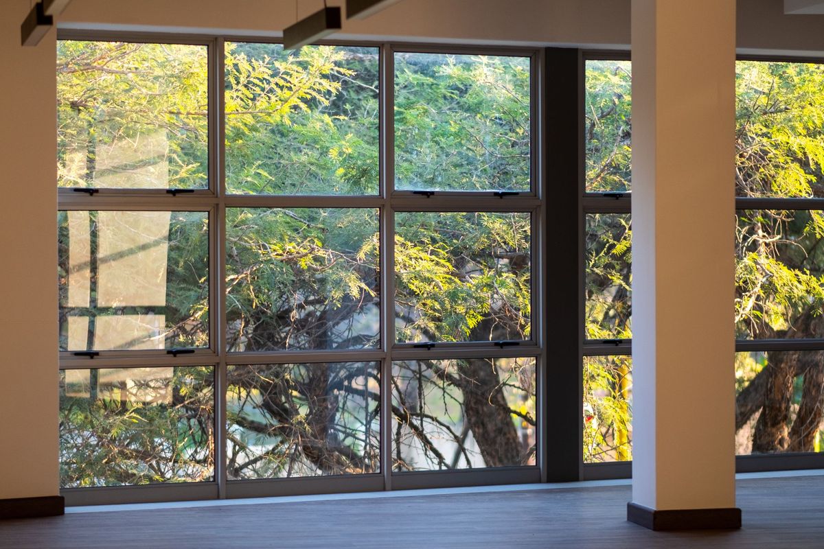A bright, open-plan office space featuring large windows with natural views, a structural column, and modern recessed track lighting.