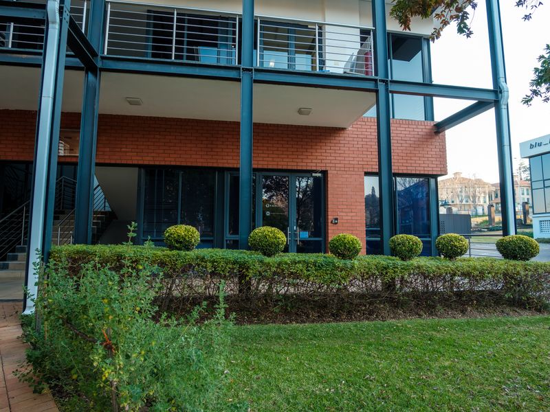 Modern office building exterior featuring a prominent exposed steel structural frame, face brick facade, and large glass windows with a landscaped garden frontage.