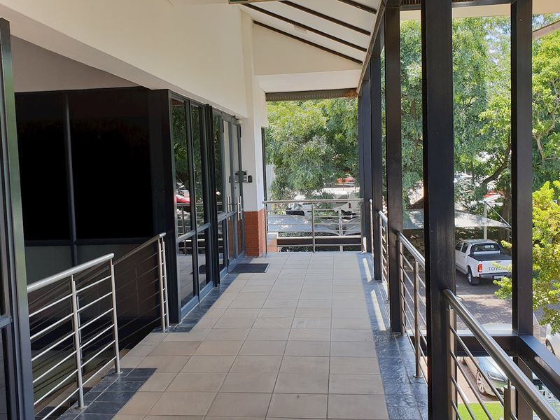 A covered outdoor balcony walkway featuring tiled flooring, steel balustrades, and large glass office frontage overlooking the parking area.