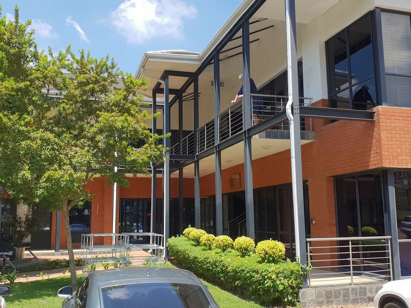 A modern office building exterior featuring red brick walls, steel balcony structures, and large glass windows overlooking a landscaped courtyard area.