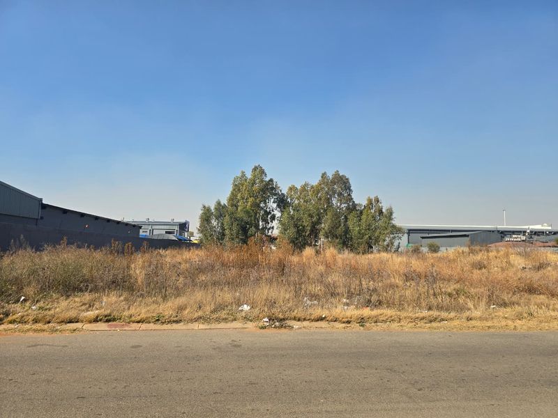 A view of a vacant commercial plot from the street, featuring overgrown grass and adjacent industrial structures.