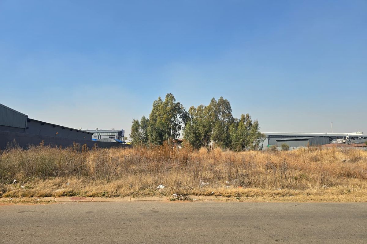 A view of a vacant commercial plot from the street, featuring overgrown grass and adjacent industrial structures.