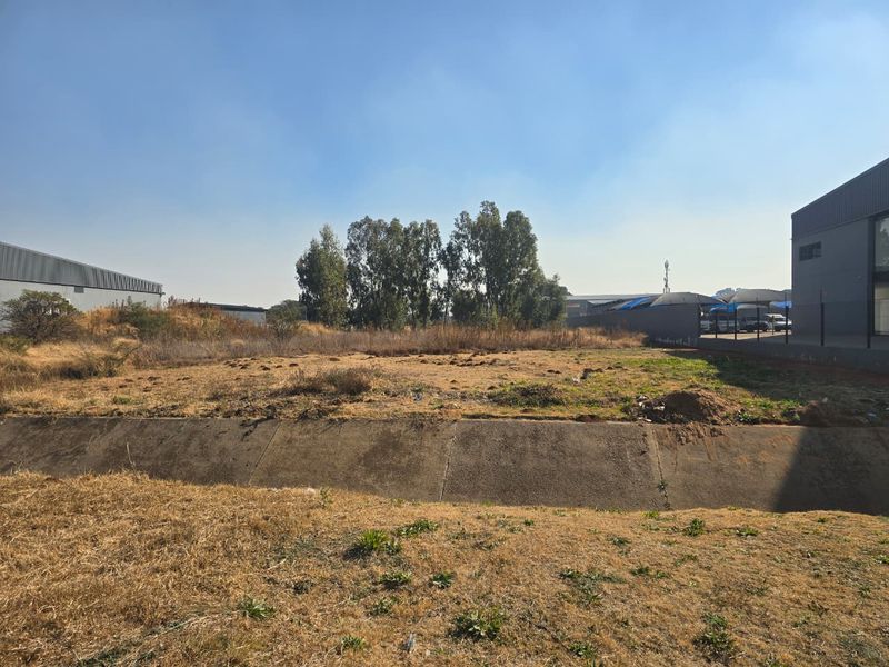 A vacant commercial land plot featuring a concrete drainage channel, dry grass, and adjacent industrial structures.