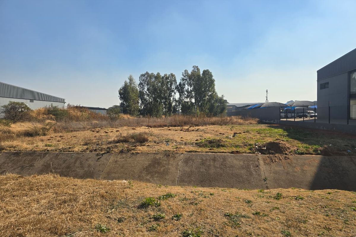 A vacant commercial land plot featuring a concrete drainage channel, dry grass, and adjacent industrial structures.
