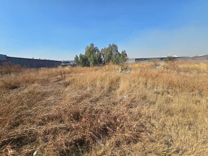 A view of an undeveloped commercial vacant land plot featuring dry, overgrown vegetation and surrounding industrial structures.