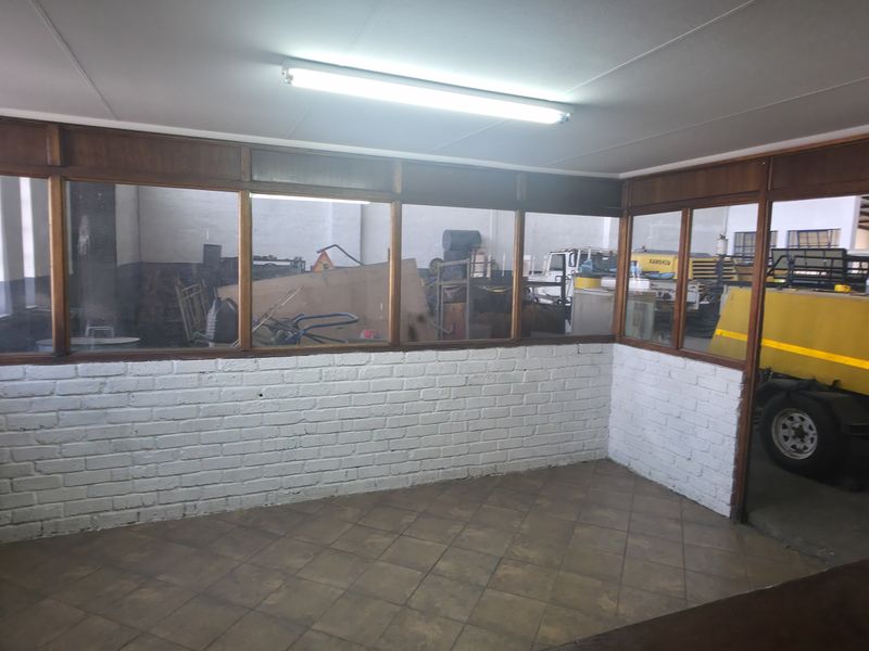 An empty office space featuring timber-framed glass partitions overlooking an industrial warehouse area, with painted brick walls and ceramic tiled flooring.