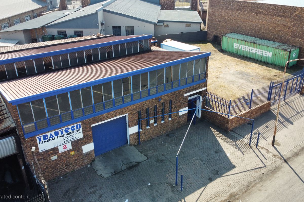 Aerial view of an industrial property featuring a brick warehouse building with a metal roof, roller shutter door, and a paved yard with perimeter fencing.