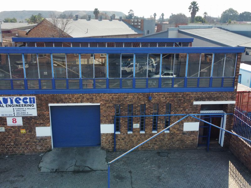 Industrial property featuring a brick facade, roller shutter door, and upper-level office glazing with a paved driveway and secure perimeter fencing.