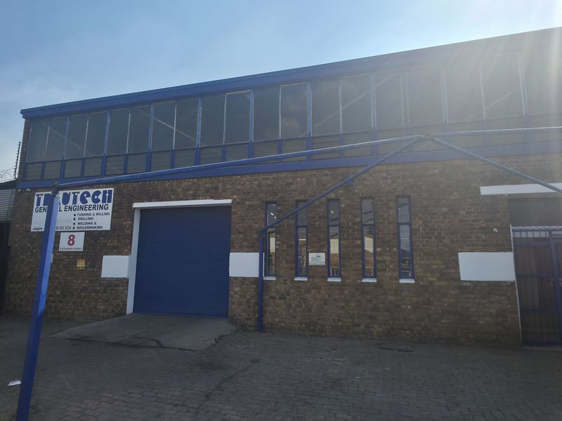 Front elevation of an industrial engineering facility featuring a brick facade, roller shutter door, and upper-level windows with security fencing.