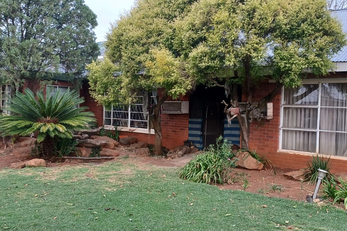 A single-story brick small holding residence featuring a corrugated metal roof, large windows, and established garden surroundings.
