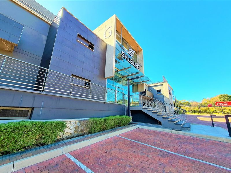 Modern office building exterior featuring a glass facade, stainless steel railings, and a paved parking area with landscaped hedging.