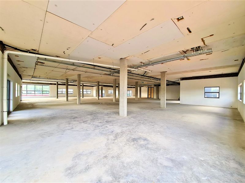 A large, open-plan office space in shell condition, featuring exposed concrete columns, unfinished ceiling panels, and expansive windows providing natural light.
