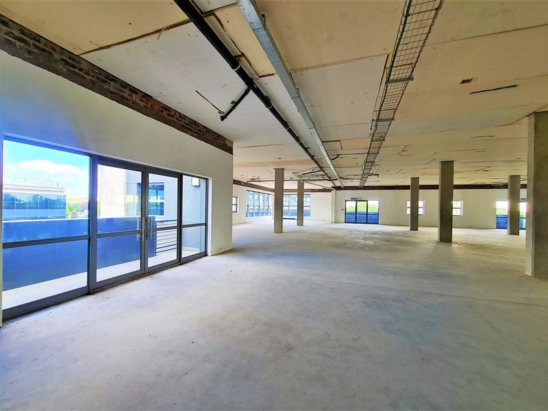 A large, open-plan office space in shell condition, featuring exposed industrial ceilings, concrete columns, and large glass doors providing natural light.