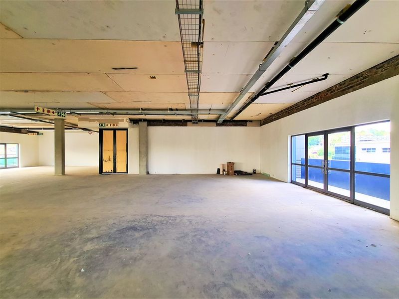 Spacious open-plan office area featuring exposed ceilings, concrete flooring, and large glass doors providing natural light and access to an exterior balcony.