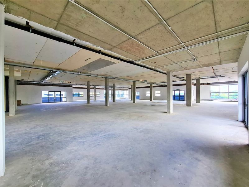 A large, open-plan office space in shell condition, featuring exposed concrete ceilings, support columns, and expansive windows providing ample natural light.