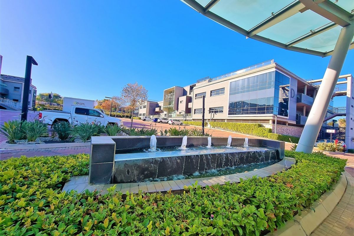 A modern commercial office complex featuring a landscaped garden, a decorative water fountain, and a glass-covered entrance canopy.