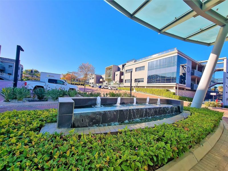 A modern commercial office complex featuring a landscaped garden, a decorative water fountain, and a glass-covered entrance canopy.