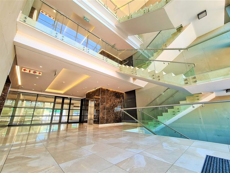 Modern office building lobby featuring a grand staircase, glass balustrades, marble wall accents, and polished porcelain flooring with recessed lighting.