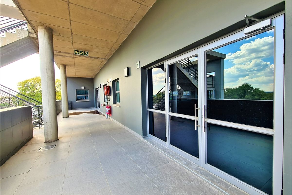 A covered exterior walkway featuring concrete columns, tiled flooring, and modern aluminium-framed glass office entrance doors.